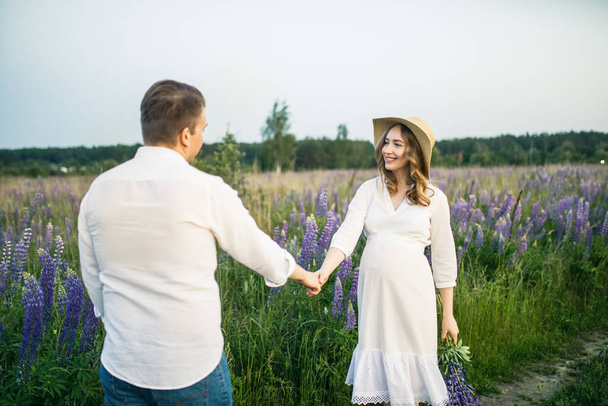 young beautiful couple a girl is pregnant in a white dress and a hat a man in a white shirt are walking along the field with lupins at sunset - Photo, image