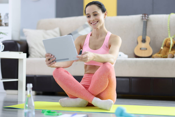 Frau in Sportbekleidung sitzt auf Gymnastikmatte und hält Tablet in der Hand - Foto, Bild
