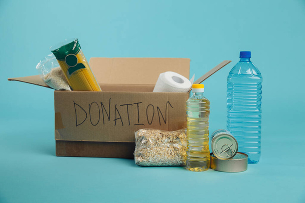 Food donations concept. Various canned food, pasta and cereals in a cardboard box on a blue background - Photo, Image