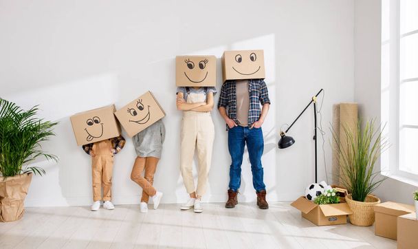 Familia irreconocible: pareja y niños con cajas de cartón en las cabezas de pie cerca de la pared en un nuevo apartamento espacioso durante la reubicación - Foto, imagen
