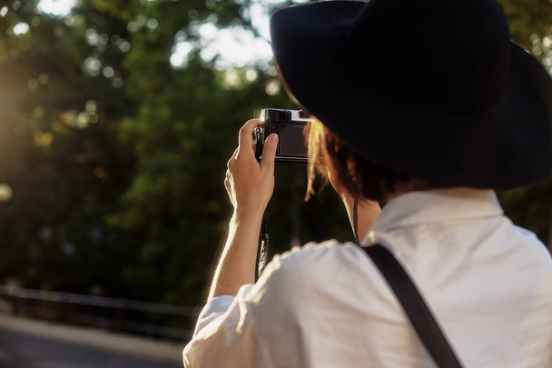 Νεαρή φωτογράφος. Το κορίτσι έχει κοντά μαύρα μαλλιά. Φοράει ένα μαύρο καπέλο και γυαλιά ηλίου με μια κάμερα να στέκεται στο δρόμο στην παλιά ευρωπαϊκή πόλη. - Φωτογραφία, εικόνα