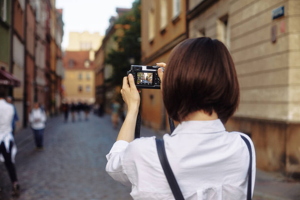 Młoda fotograf. Dziewczyna ma krótkie czarne włosy. Ma na sobie czarny kapelusz i okulary przeciwsłoneczne z kamerą stojącą na drodze w starym europejskim mieście. - Zdjęcie, obraz