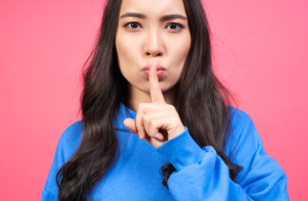 Serious mysterious brunette Asian woman presses index finger to lips makes hush gesture tells secret asks to be quiet, poses against pink background. Secrecy concept. - Foto, imagen