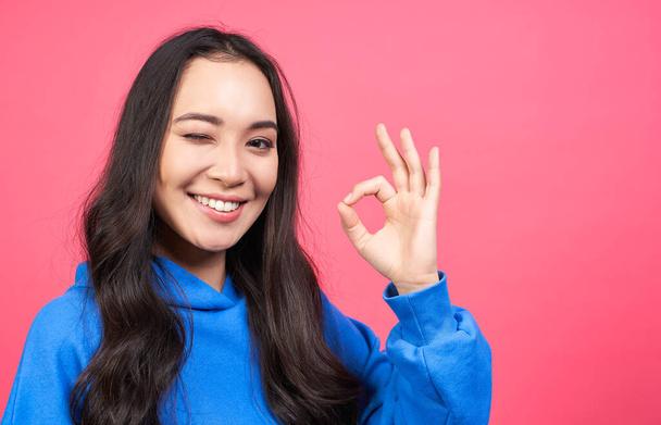 Photo of cheerful young woman blinks eye, makes okay gesture, demonstrates her agreement, feels happy, isolated over pink background. Body language concept. Copy space. - Photo, image