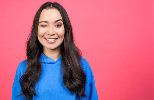Photo of cheerful young woman blinks eye, wears blue sweatshirt, demonstrates her agreement, feels happy, isolated over pink background. Body language concept. Copy space. - Foto, Bild