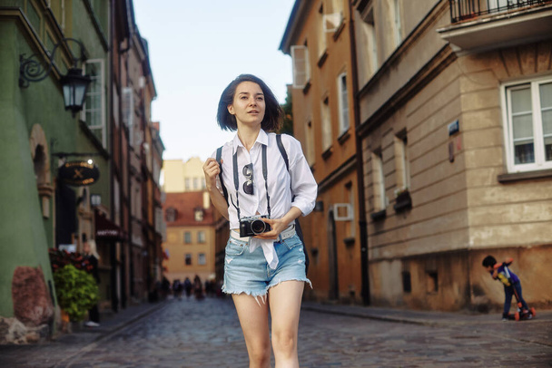 Young girl-photographer. The girl has short black hair. She is wearing a black hat and sunglasses with a camera standing on the road in the old European city. - Foto, afbeelding