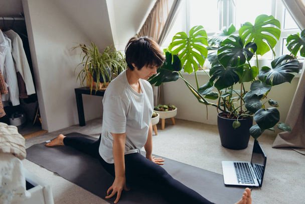 A woman on a mat trying to do a twine asana position in her bedroom with plants. Online class yoga practice at home. Healthy Lifestyle. Home online workout during a lockdown. Soft selective focus. - Foto, Imagem