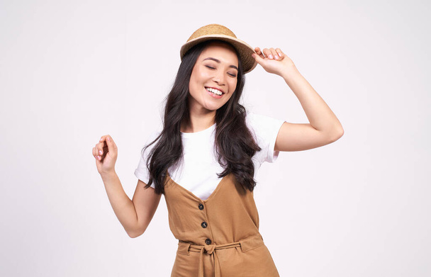Cute smoldering pretty Asian girl with long black hair in straw hat dancing and having fun, holding her hands up, wearing a white t-shirt, moving to music, closing her eyes with joy, white background. - Фото, зображення