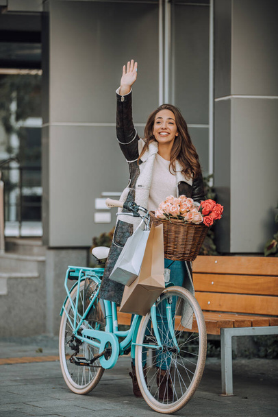 Hermosa joven sonriente mujer caminando con bicicleta, después de ir de compras y saludar la mano de alguien, en la calle de la ciudad. Belleza, gesto de saludo y concepto de estilo de vida - Foto, imagen