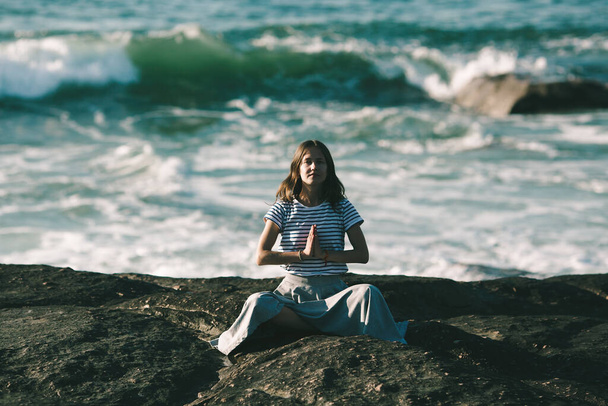 Piękna młoda kobieta jogi medytuje w pozycji lotosu, siedząc na skałach na brzegu oceanu. - Zdjęcie, obraz
