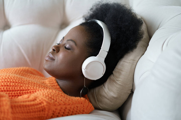 Μαύρη γυναίκα με πορτοκαλί πουλόβερ που φοράει ασύρματα ακουστικά, ακούει μουσική, ξεκουράζεται, ξαπλωμένη στον καναπέ - Φωτογραφία, εικόνα