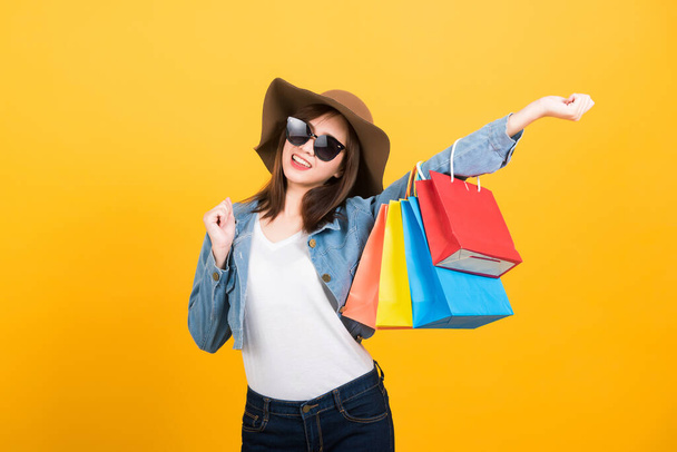 Asian happy portrait beautiful cute young woman teen smiling standing with sunglasses excited holding shopping bags multi color looking camera isolated, studio shot yellow background with copy space - Photo, image