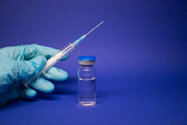 Разработка коронавирусной вакцины COVID-19. медицинский шприц с иглой для инъекций в синей медицинской перчатке рядом с бутылкой, без этикетки. изолированы на синем фоне. Лечение. копировальное пространство - Фото, изображение
