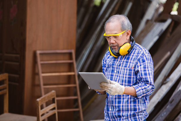 Asyalı kıdemli marangoz iş yerinde duran tableti kullanarak kulaklık takıyor. marangoz konsepti. - Fotoğraf, Görsel