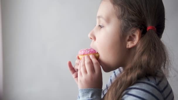Kis kaukázusi lány rózsaszín fánkot eszik. Élvezem. Közelről.. - Felvétel, videó
