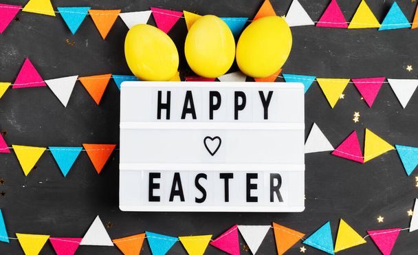 Happy easter lettering on white board with felt garland and yellow painted eggs on dark background - Фото, зображення