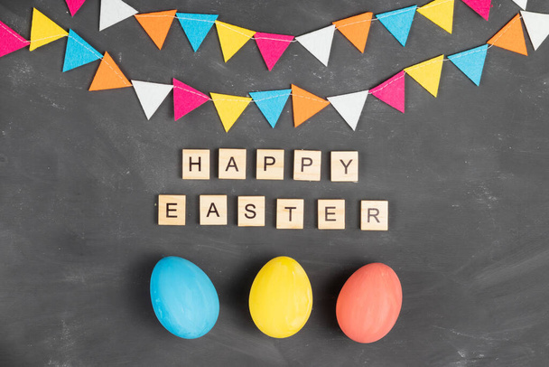Happy easter lettering on wooden squares on chalk board with painted eggs and colorful felt garland - Фото, изображение