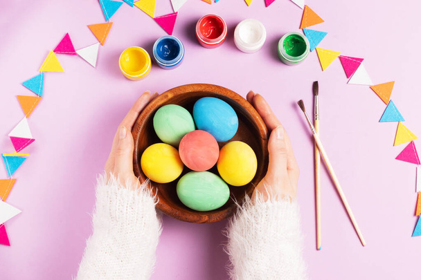 Female hands in a white fluffy sweater hold a wooden plate with colorful easter eggs on a pink background with paints, brushes and a garland of felt - Foto, Bild