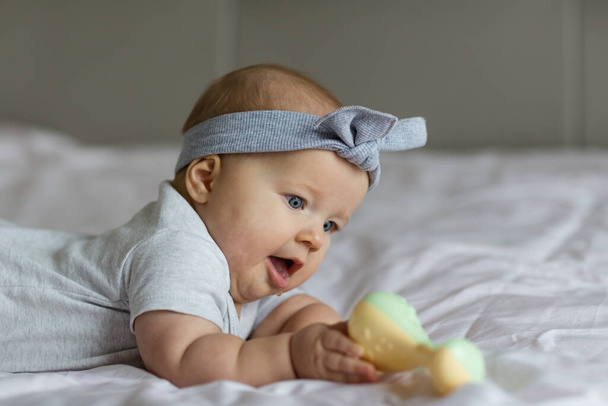 Caucasian blonde baby six months old lying on bed at home and playing with rattle toy. Kid wearing cute gray clothing. Bedroom decorated with flags. - Photo, image