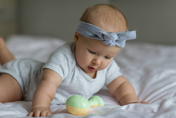 Caucasian blonde baby six months old lying on bed at home and playing with rattle toy. Kid wearing cute gray clothing. Bedroom decorated with flags. - Photo, image