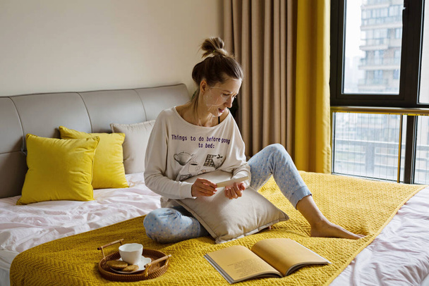 Millennial Caucasian woman with blonde hair relaxing on bed at home during coronavirus Covid-19 pandemic quarantine - Valokuva, kuva