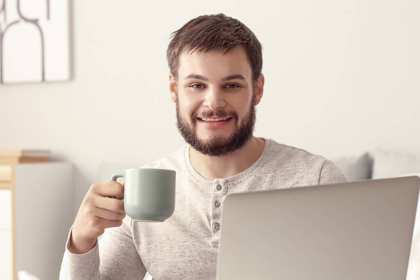 Young man with laptop and cup of tea in bedroom - Φωτογραφία, εικόνα