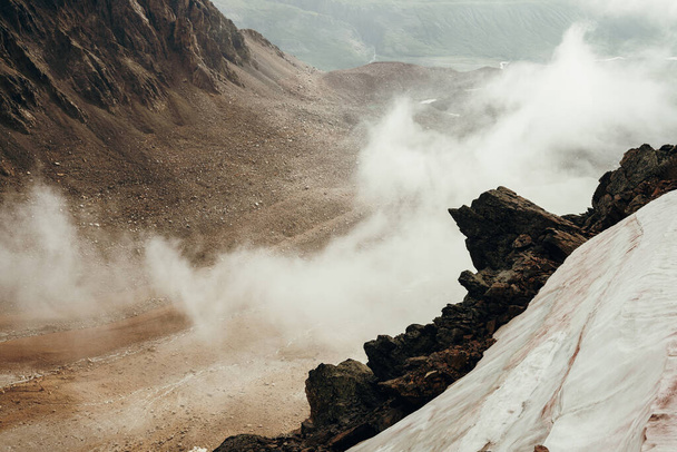 Όμορφος μυτερός γκρεμός στην ψηλή βραχώδη άκρη του βουνού με χιόνι ανάμεσα σε πυκνά χαμηλά σύννεφα. Ατμοσφαιρικό μινιμαλιστικό αλπικό τοπίο Σημαδεμένη πέτρα κοντά στην άβυσσο στα σύννεφα. Υπέροχο ορεινό τοπίο. - Φωτογραφία, εικόνα