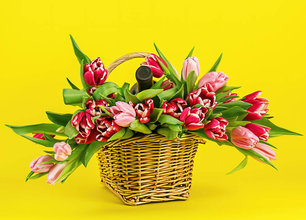 Basket with colorful bouquets of tulips and bottle of wine on yellow - Photo, Image