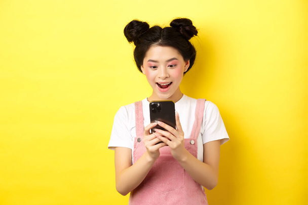Прелестная девочка, взволнованно смотрящая на экран, читающая сообщения по телефону и счастливо улыбающаяся, стоящая в летней одежде на желтом фоне - Фото, изображение
