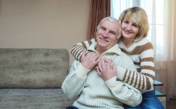 A married couple of 50-60 years old are hugging while sitting on the sofa at home. Looking into the camera. The concept of a long happy marriage. Place for text. - Fotó, kép