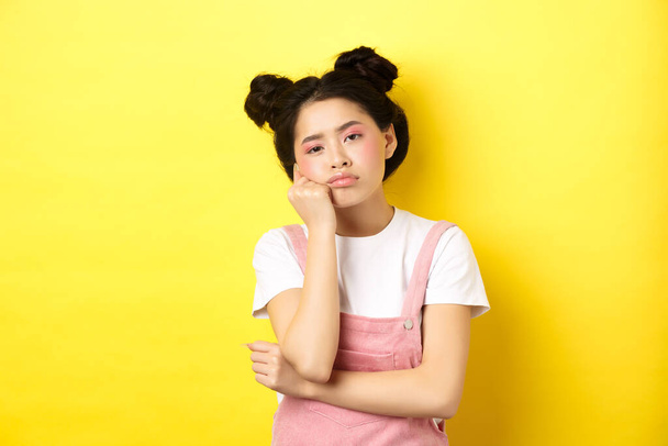 Sad and bored teen asian girl standing alone, looking at camera indifferent, staring with boredom, yellow background - Foto, afbeelding