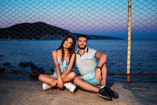 Un jeune couple est assis près de la clôture sur le béton, contre la mer. Au crépuscule. Le concept de Saint-Valentin. - Photo, image