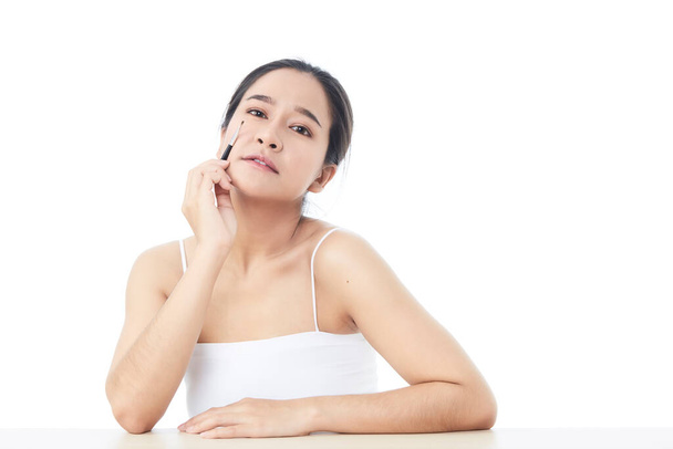 Beautiful Asian woman looking at the camera and with a small cosmetics brush in her raised a hand for contouring her eyebrow headshot with bare shoulders on white background - Photo, Image
