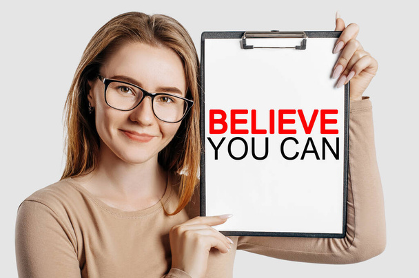 Believe you can. Beautiful young business woman wearing glasses holds a clipboard with mock up space isolated on gray background - Fotoğraf, Görsel