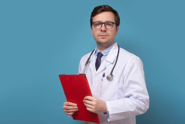 Dojrzały lekarz stojący z folderem na papierze na niebieskiej ścianie. - Zdjęcie, obraz