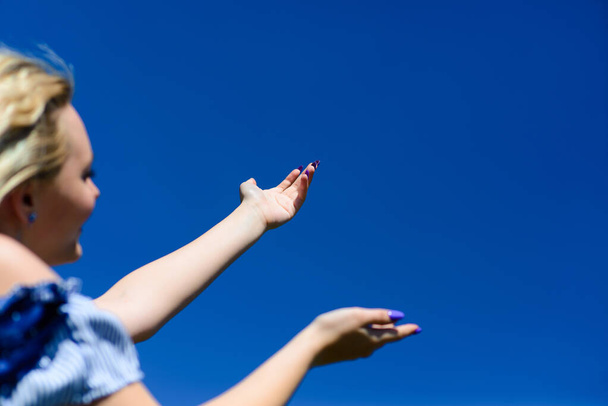 Μια νεαρή ξανθιά κοπέλα κοιτάζει τον όμορφο γαλάζιο ουρανό και φτάνει στον ουρανό με τα χέρια της. Όμορφη γυναίκα θαυμάζει τη φύση. - Φωτογραφία, εικόνα