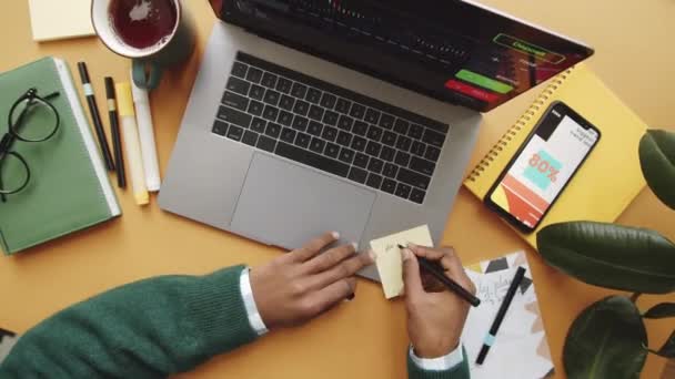 Time lapse top down shot van handen van onherkenbare Afro-Amerikaanse man typen op laptop, met behulp van smartphone, notities maken en thee drinken tijdens het werken op kantoor - Video