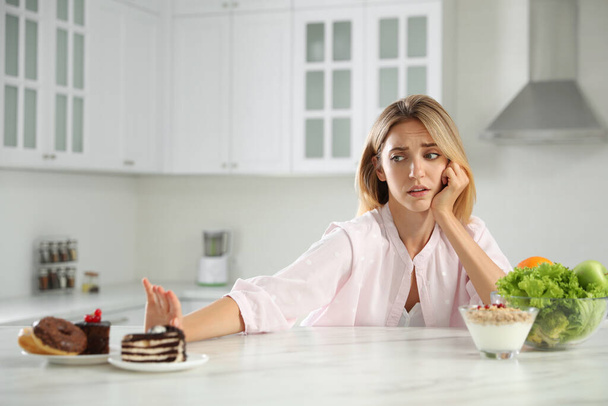 Woman choosing between sweets and healthy food at white table in kitchen - Foto, Bild