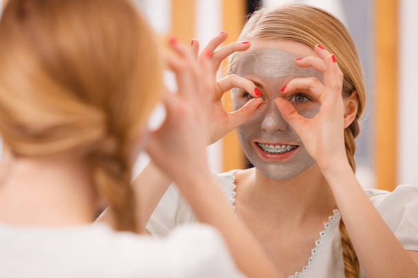 Skincare. Young positive woman with grey clay mud mask on her face having fun. Teenage girl taking care of skin condition, folded her hands as glasses. Spa beauty treatment. - Foto, Bild