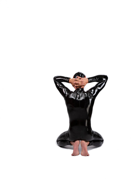 Visão traseira retrato de um escravo sentado e segurando as mãos atrás da cabeça de joelhos, imagem de estúdio em fundo branco isolado. Homem vestido com roupas de fetiche de borracha de látex na moda  - Foto, Imagem