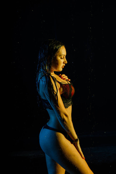 Frau im weißen Hemd mit Wassertropfen in einem dunklen Raum, der von Licht beleuchtet wird, während eines Fotoshootings mit Wasser in einem kleinen Pool - Foto, Bild