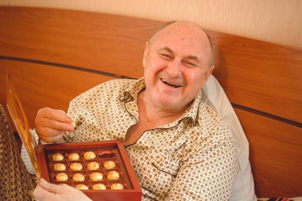 An elderly happy man in pajamas has chocolate sweets in bed in the morning. Psychology of mature men, getting rid of depression and bad mood - Photo, Image