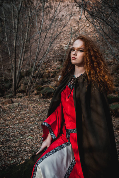 Red-haired woman in a red dress in a historical Celtic costume in the autumn forest - Photo, Image