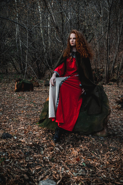 Red-haired woman in a red dress in a historical Celtic costume in the autumn forest - Foto, Imagen