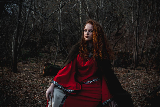 Red-haired woman in a red dress in a historical Celtic costume in the autumn forest - Fotoğraf, Görsel