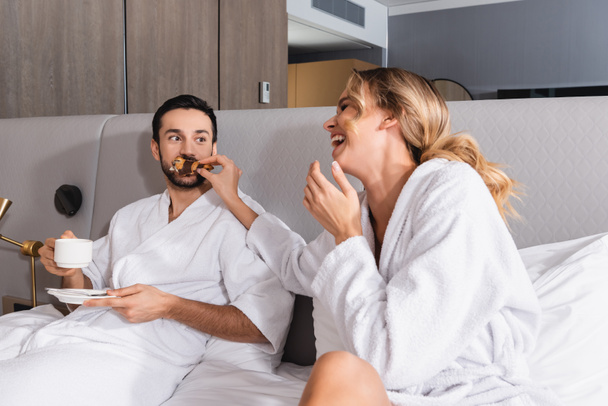 Cheerful woman feeding croissant to muslim boyfriend with coffee on hotel bed  - 写真・画像