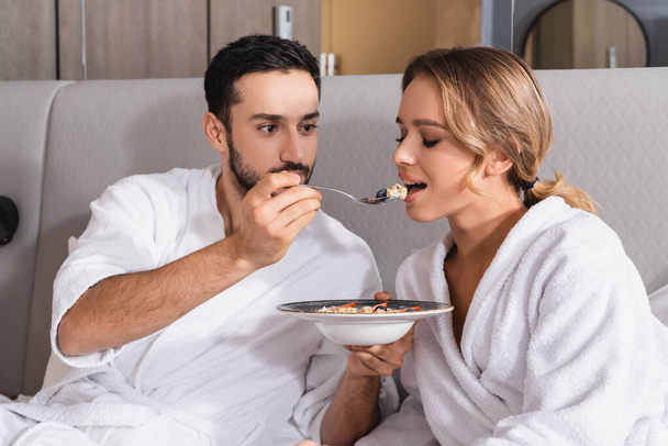 Arabian man in bathrobe feeding salad to girlfriend on hotel bed  - Foto, Imagen