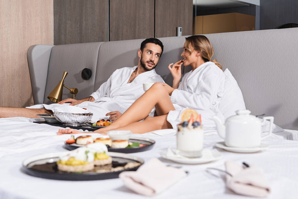 若いです異人種間のカップルでバスローブ座っています近くにおいしい食べ物上のぼやけたフォアグラウンドオンホテルベッド  - 写真・画像