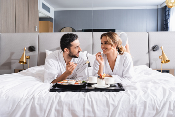 Uśmiechnięta międzyrasowa para patrząca na siebie przy śniadaniu na tacy na łóżku hotelowym  - Zdjęcie, obraz