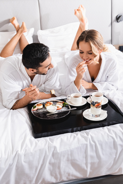 Uśmiechnięta kobieta patrząca na desery na tacy obok muzułmańskiego chłopaka na łóżku hotelowym  - Zdjęcie, obraz